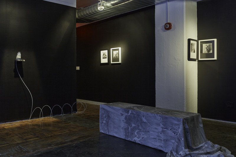 Facing the other direction in the same room; four black and white prints are hung on a wall painted black. On the left wall is a teacup containing a blue liquid with foam pouring out the top; a tube spirals to the floor and out of sight.